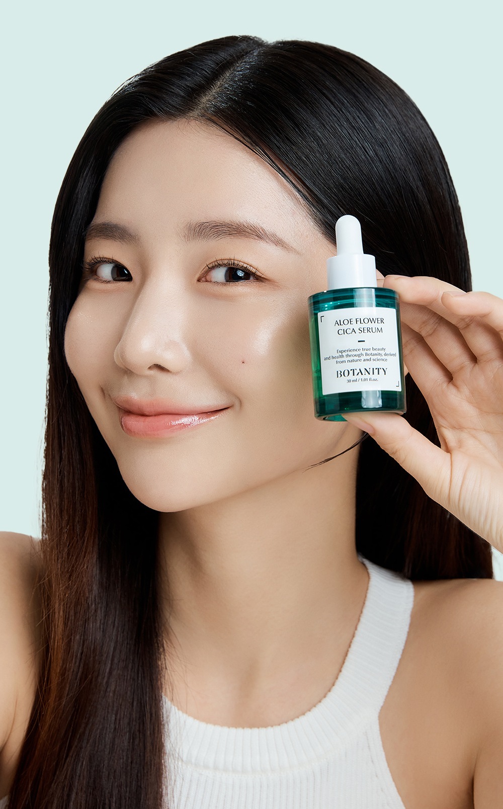 모델이 뺨 옆에 보타니티 알로에 플라워 시카 세럼 병을 들고 미소 짓는 사진, 민트 벽. 병 글자: ALOE FLOWER CICA SERUM, BOTANITY, 30 ml / 1.01 fl.oz.