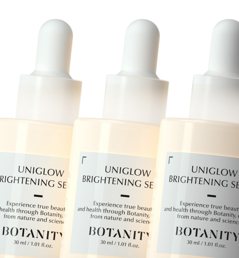 흰 스포이트 세럼 병 셋이 비스듬히 나란히 선 근접 사진 — 뒤에서 비치는 따뜻한 빛. 라벨 UNIGLOW BRIGHTENING SERUM, BOTANITY, 30 ml / 1.01 fl.oz.